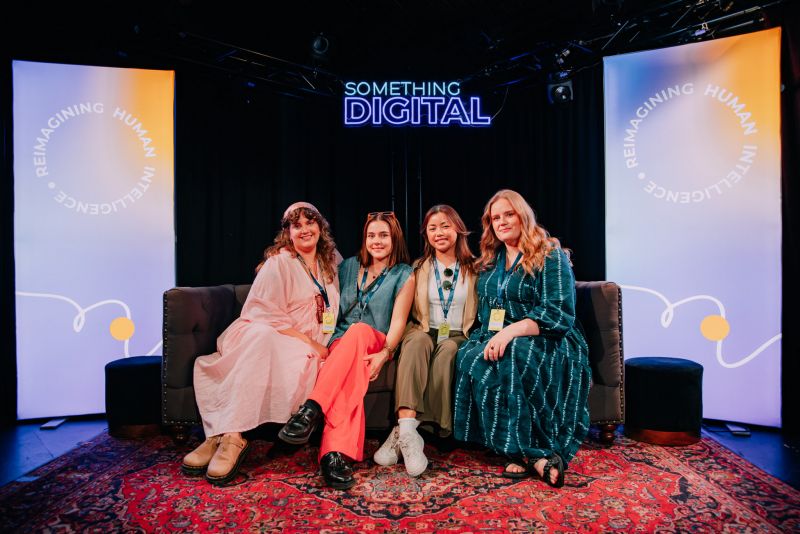 A photo of four women sitting on a dark grey couch. The setting appears to be a stage, with two vertical banners on either side of the couch that read, 