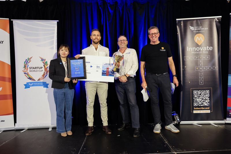 Four people stand on a stage during a Startup World Cup event, holding a winner’s plaque, trophy and oversized prize cheque. Event banners for Startup World Cup and Innovate Moreton Bay are displayed on either side against a blue-lit backdrop.