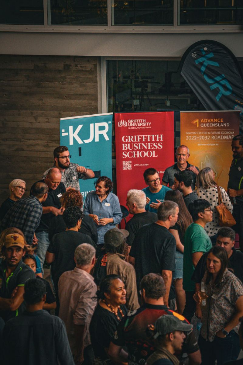 A diverse group of people, including men and women of various ages, are gathered in a crowded indoor space. They are casually dressed and are engaged in conversation, with some holding drinks. In the background, there are three large banners.