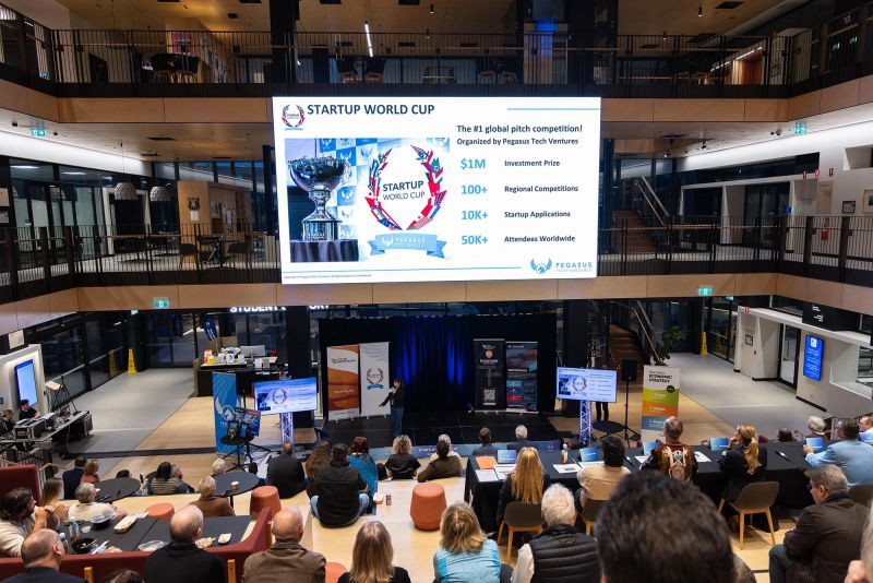 2.	Audience members watch a Startup World Cup pitch event in a large indoor atrium. A presenter speaks on stage beneath a large digital screen displaying Startup World Cup competition information, while attendees are seated in tiered viewing areas around the venue.