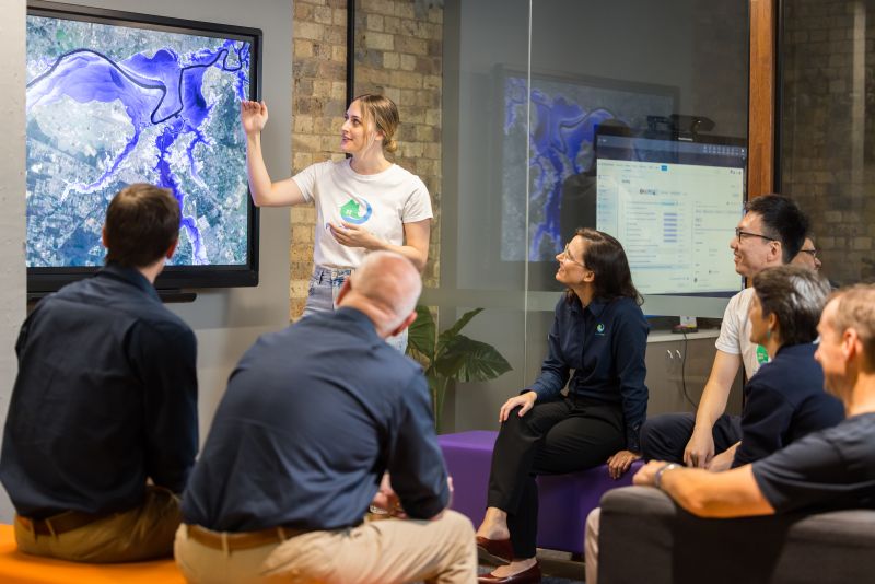 A woman presenting on a screen to an audience of people seated casually around here.
