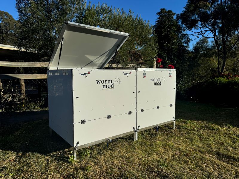 A photo of a white, rectangular modular worm farm system, labeled 'worm mod' on each section, sits outdoors on green grass. The lid of one section is open, revealing the interior. Trees and a wooden fence are visible in the background.