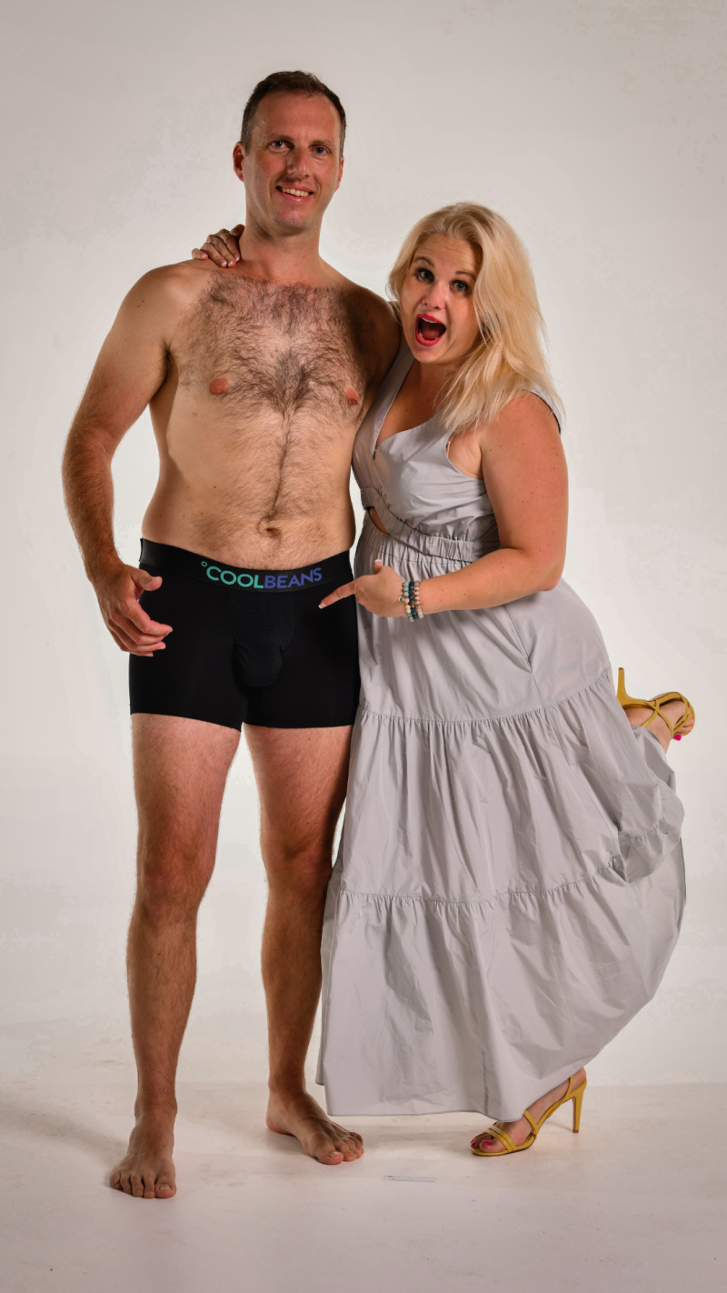 A full-length shot of a man and a woman standing together against a light background. The man is wearing black boxer briefs with the text 'COOLBEANS' visible on the waistband. He is shirtless and has his arm around the woman's shoulder. The woman is wearing a light grey dress and yellow heels, and is pointing towards the man's underwear with a surprised expression.