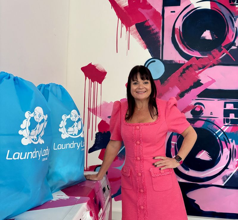 A photo portrait of CEO of Laundry Lady, Susan Toft, in a pink dress, standing beside two large, bright blue branded laundry bags. She is in front of a modern graffiti mural of stylized pink and black washing machines.