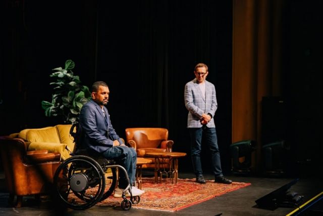 A photo shows a man in a wheelchair and one standing up on a stage.