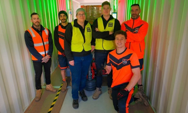 Photo of six workers inside what appears to be a brightly lit storage container or narrow hallway. They are all wearing work-appropriate attire, including high-visibility vests or jackets in orange and yellow, and some have long-sleeved shirts underneath.