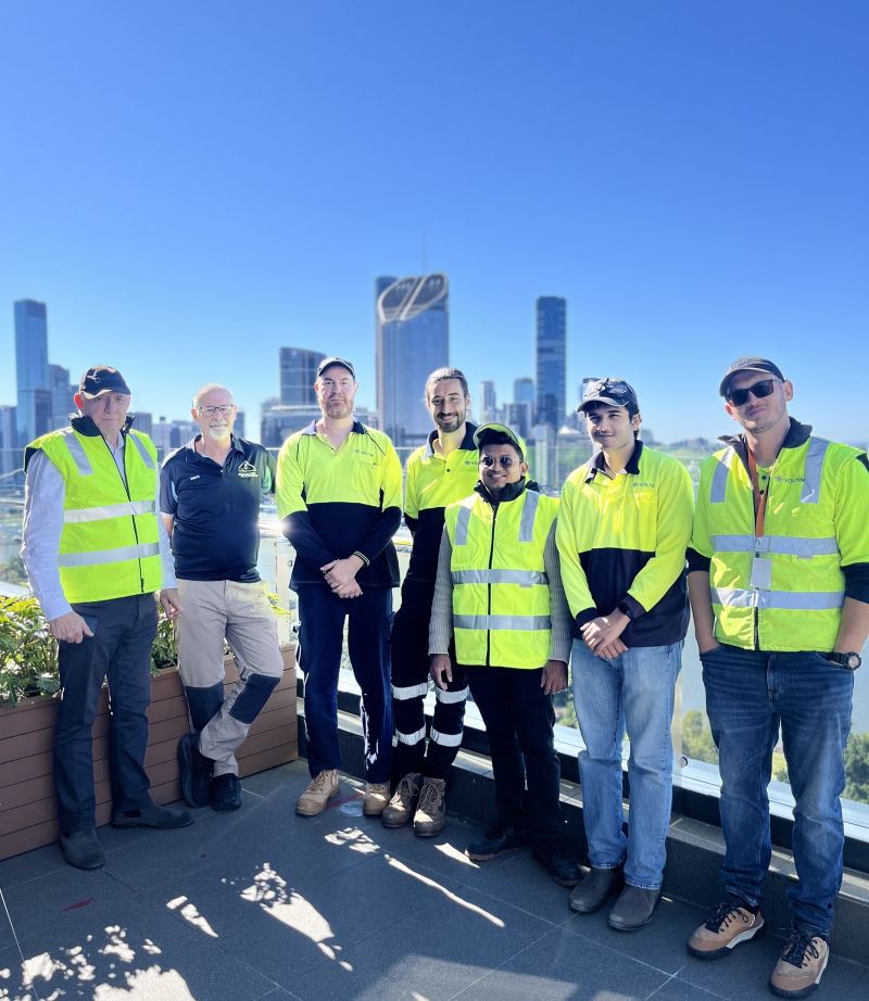 A team of seven individuals from the Voltin team stand on a rooftop deck with the skyline of Brisbane in the background. Most of the men are wearing high-visibility yellow and black vests or long-sleeve shirts.