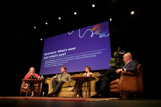 A photo shows four panelists sitting on a stage in armchairs during a panel discussion. 