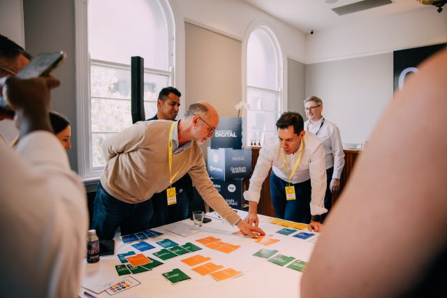 A photo shows a group of people standing around a table, engaged in a discussion. On the table are numerous blue, green, and orange cards, which some of the attendees are moving around.