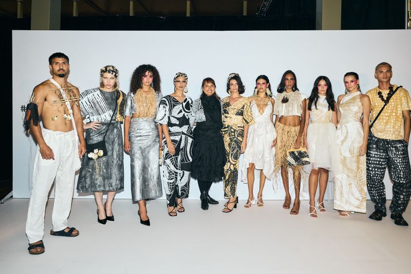 Ten models wearing a variety of artistic, avant-garde outfits stand in a row in front of a plain white background.