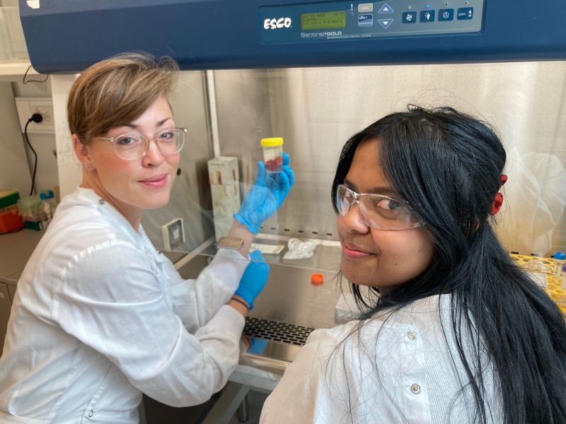 Two women, one with short brown hair and glasses, the other with long black hair, wearing lab coats and safety glasses, holding a small vial with a specimen in a lab setting.