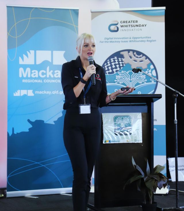 A woman stands at a podium holding a microphone in front of two banners. The banner on the left is blue and white with the 