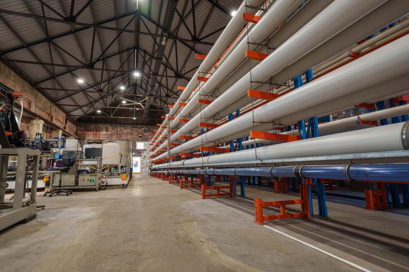 An interior view of a spacious warehouse with a high ceiling and metal beams, showing numerous long, white and blue pipes neatly stacked on multiple levels of orange and blue industrial shelving. Equipment and machinery are visible on the left side of the frame.