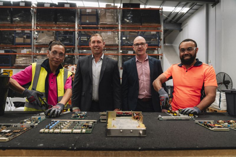 Photo of four people, two in suits and two in workwear, stand behind a table with circuit boards