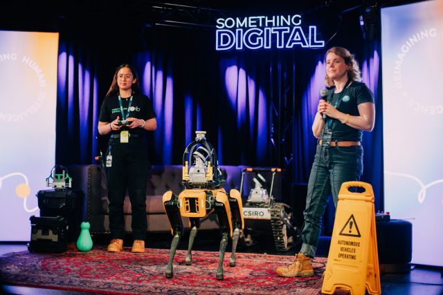 A photo shows two women on a stage, with a robot positioned between them. The robot is yellow and black and is a Boston Dynamics Spot robot with a CSIRO logo on it. 